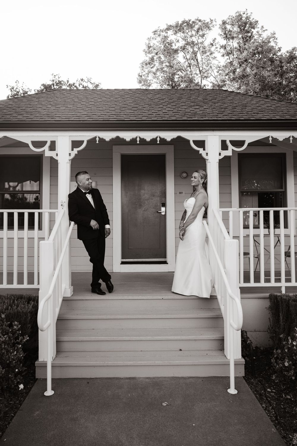 A couple in wedding attire stand on opposite sides of a charming white porch with decorative trim and railings.