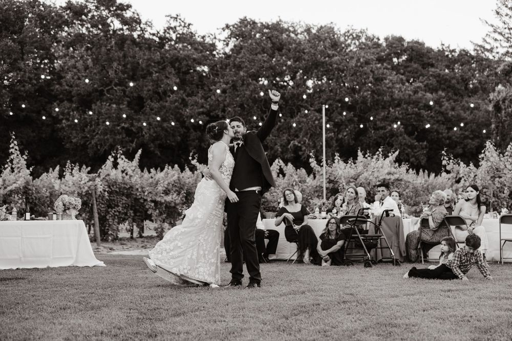 A couple shares their first dance at an outdoor garden wedding reception as guests watch from their tables in black and white.