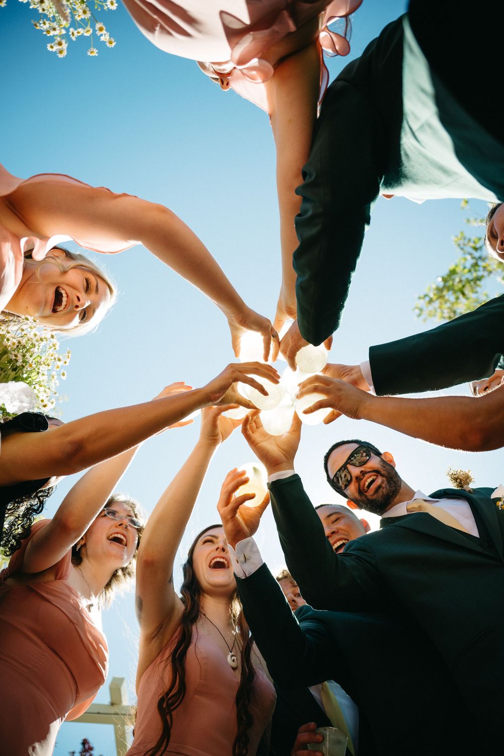 Wedding party laughing and raising hands together in a joyful circle outdoors under a bright blue sky.