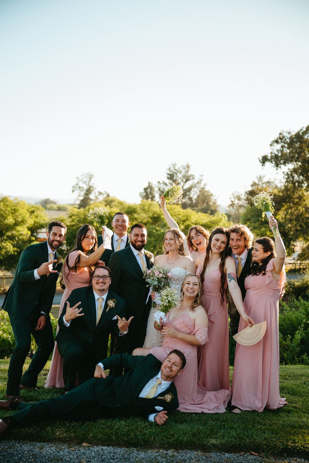 Happy wedding party posing playfully outdoors with bridesmaids in pink dresses and groomsmen in dark green suits.