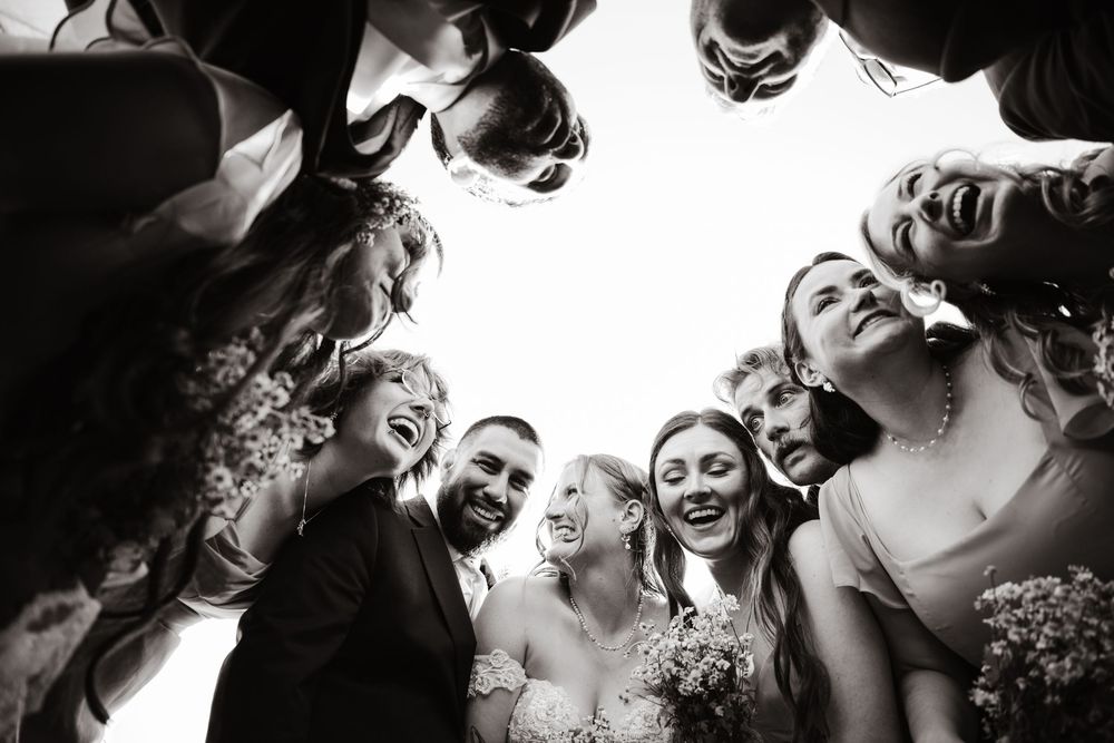 Black and white photo of wedding party huddled together laughing, shot from below looking up at smiling bride and groom.