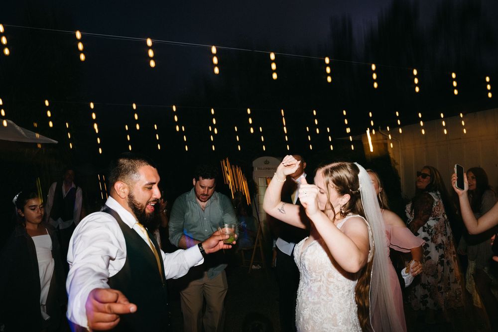 Bride and groom dancing joyfully at outdoor wedding reception under string lights at night.