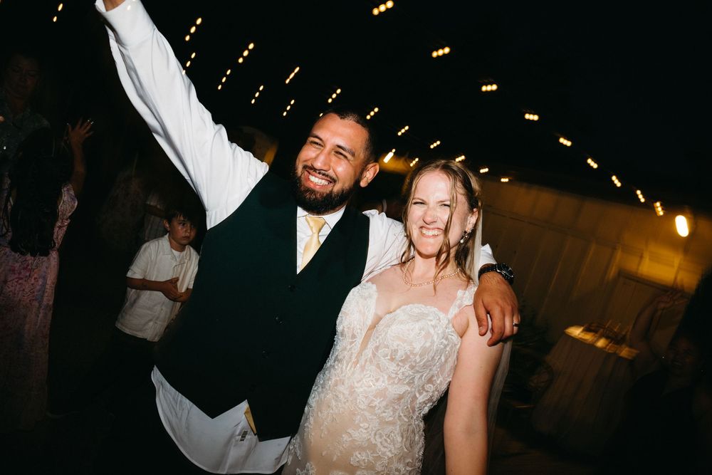 Joyful bride and groom celebrating at wedding reception, groom raising arm in excitement, string lights in background.
