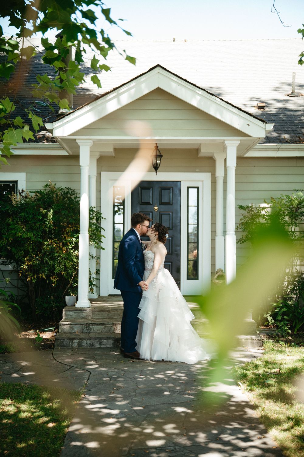 Bride and groom share a romantic moment in front of a charming white colonial home entrance on their wedding day.