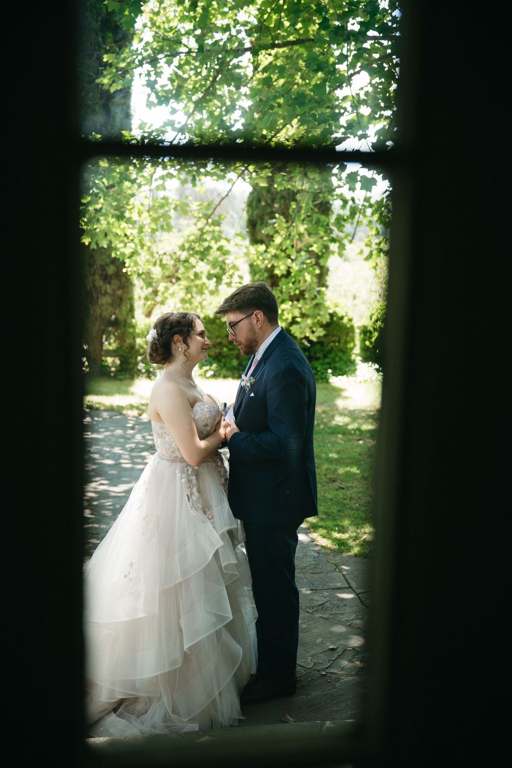 Bride and groom share a romantic moment in a lush garden, viewed through a dark doorframe on their wedding day.