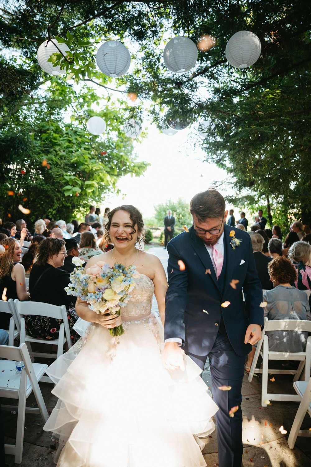 Joyful newlyweds walk down outdoor aisle as guests toss flower petals, surrounded by trees and paper lanterns.