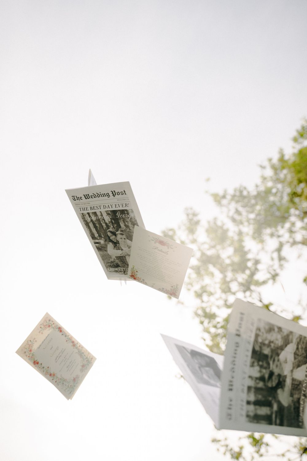 Wedding programs and menus tossed in the air against a bright sky with green trees in the background.