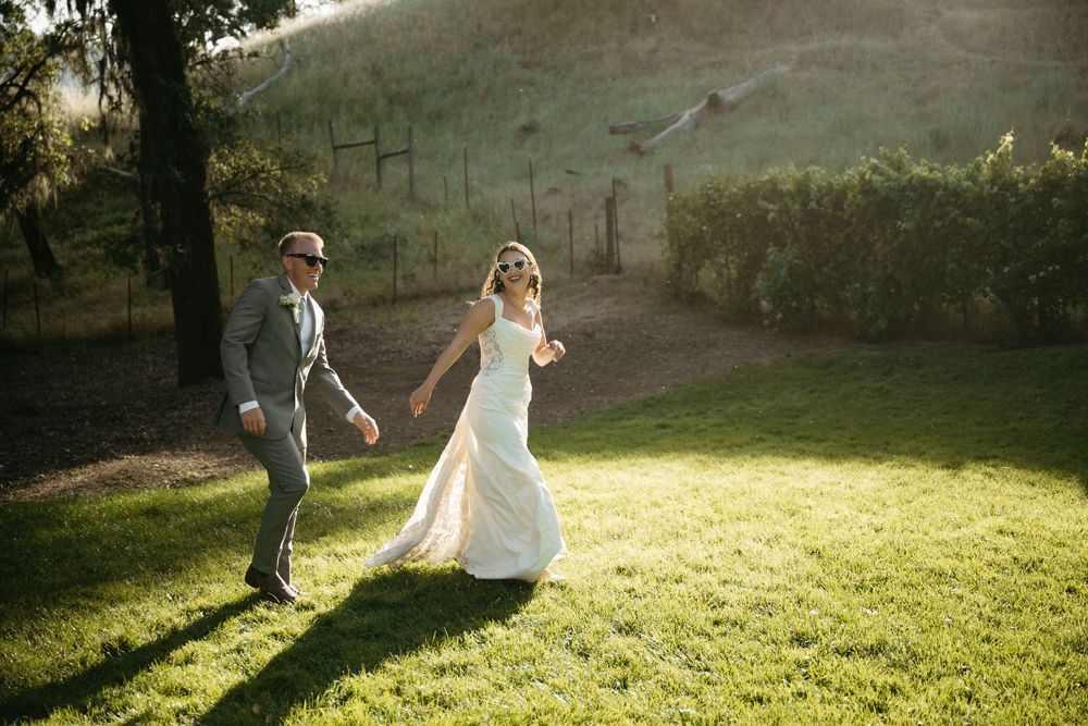 Bride and groom joyfully running hand in hand across lush green lawn during golden hour wedding celebration.