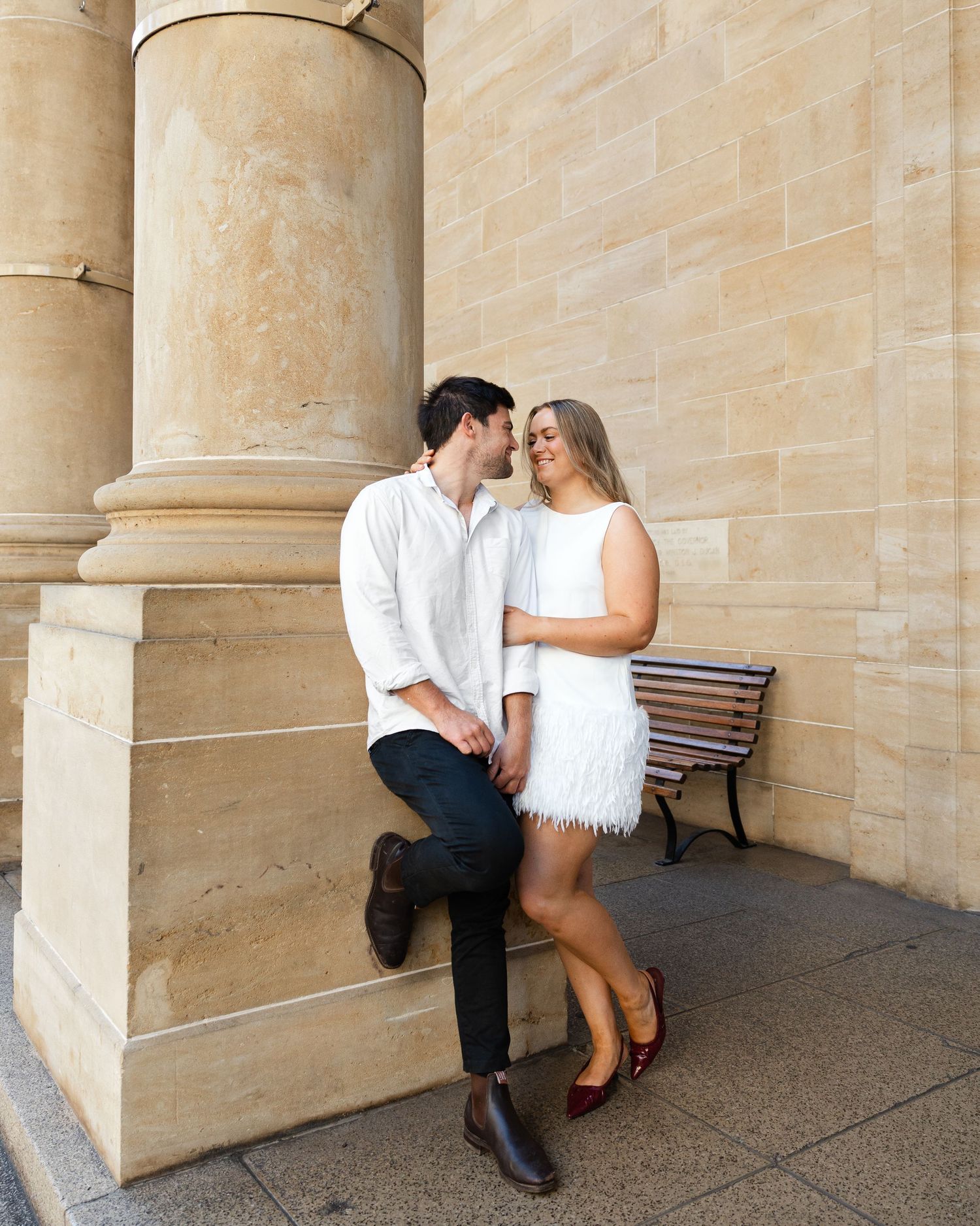 A couple shares a romantic moment while leaning against sandstone columns with a wooden bench nearby.