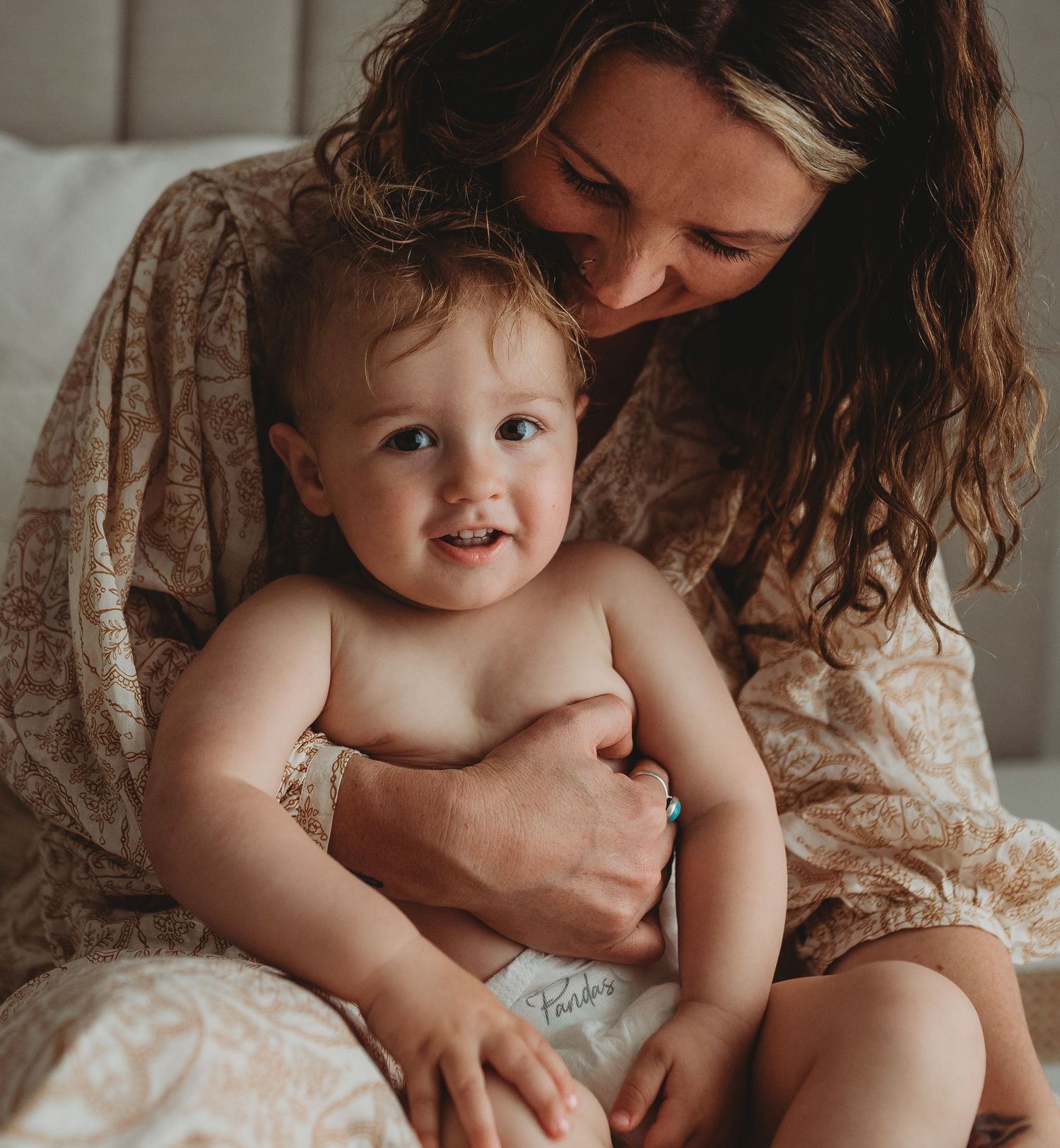 Candid lifestyle portrait shows playful interaction on white bedding in natural light.