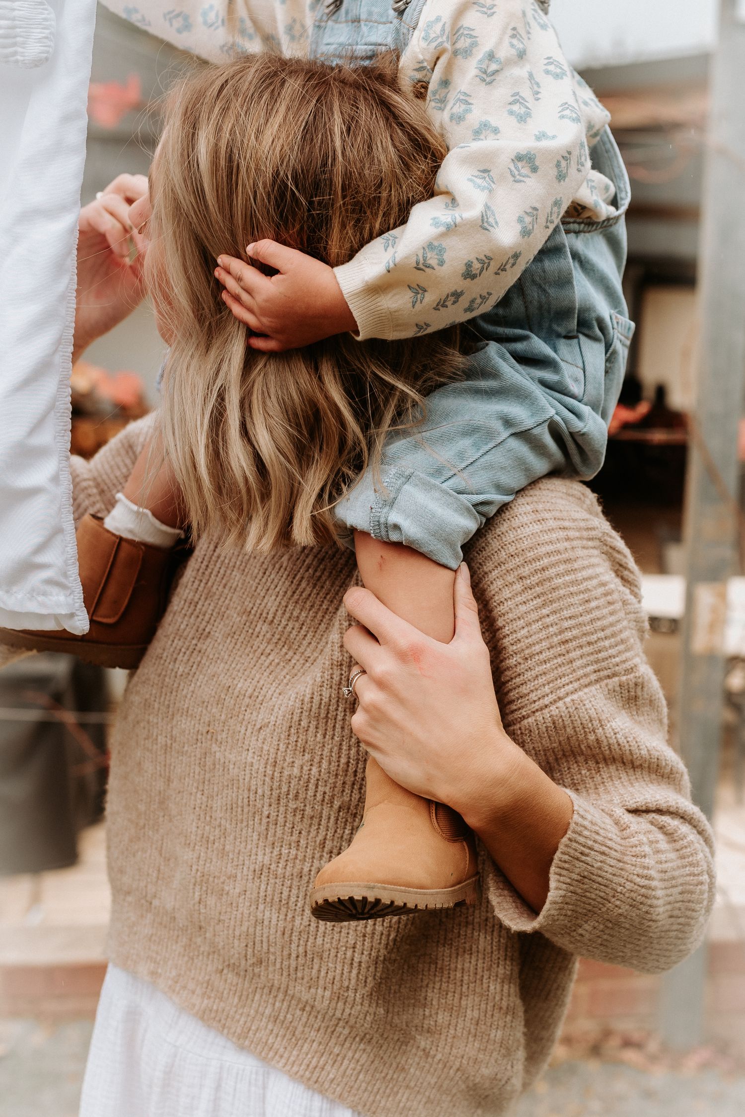 Someone wearing a beige sweater holds a child in denim overalls.