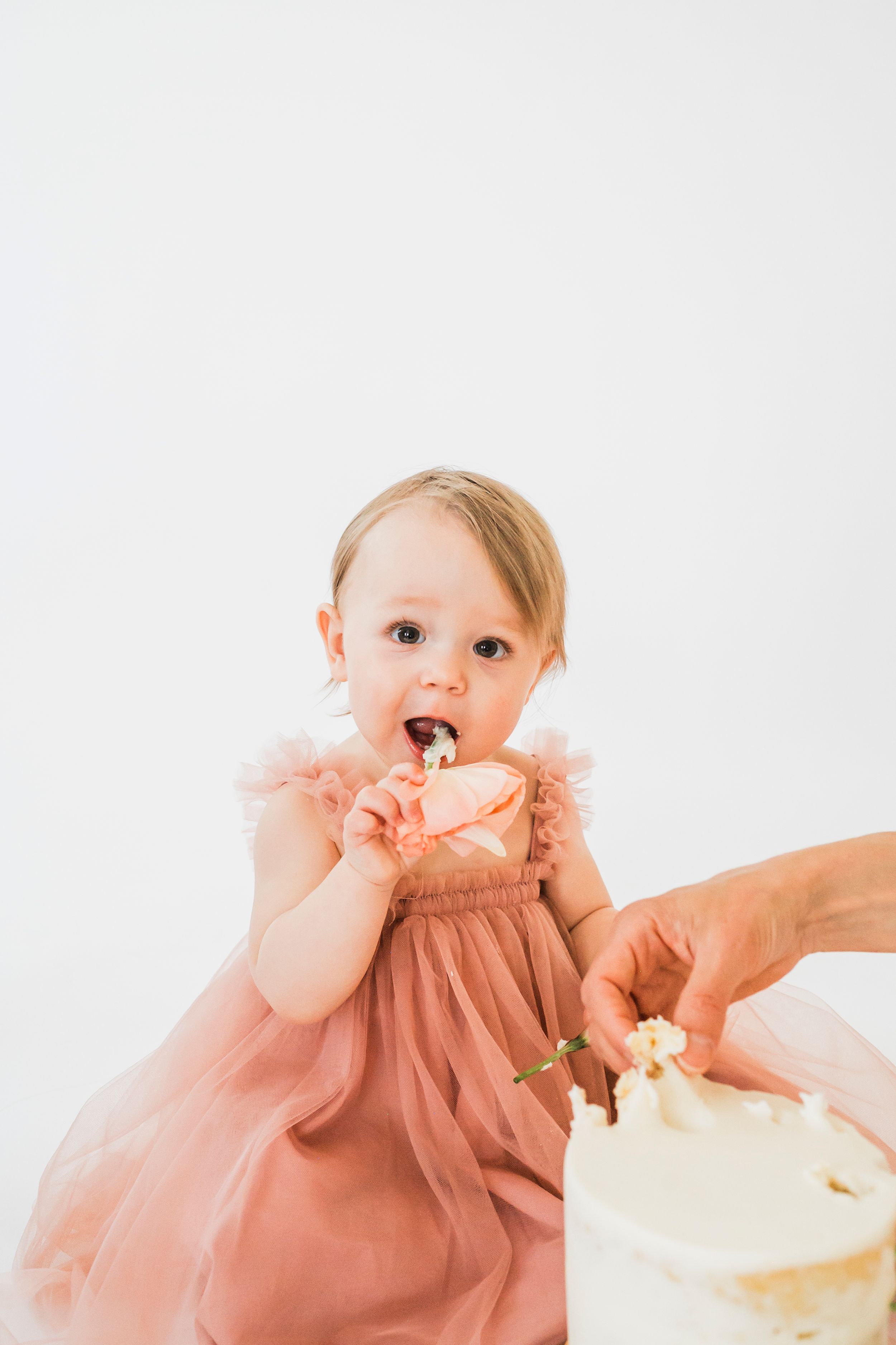 Small child in a peach-colored tulle dress enjoying their first taste of birthday cake at a cake smash photo session.