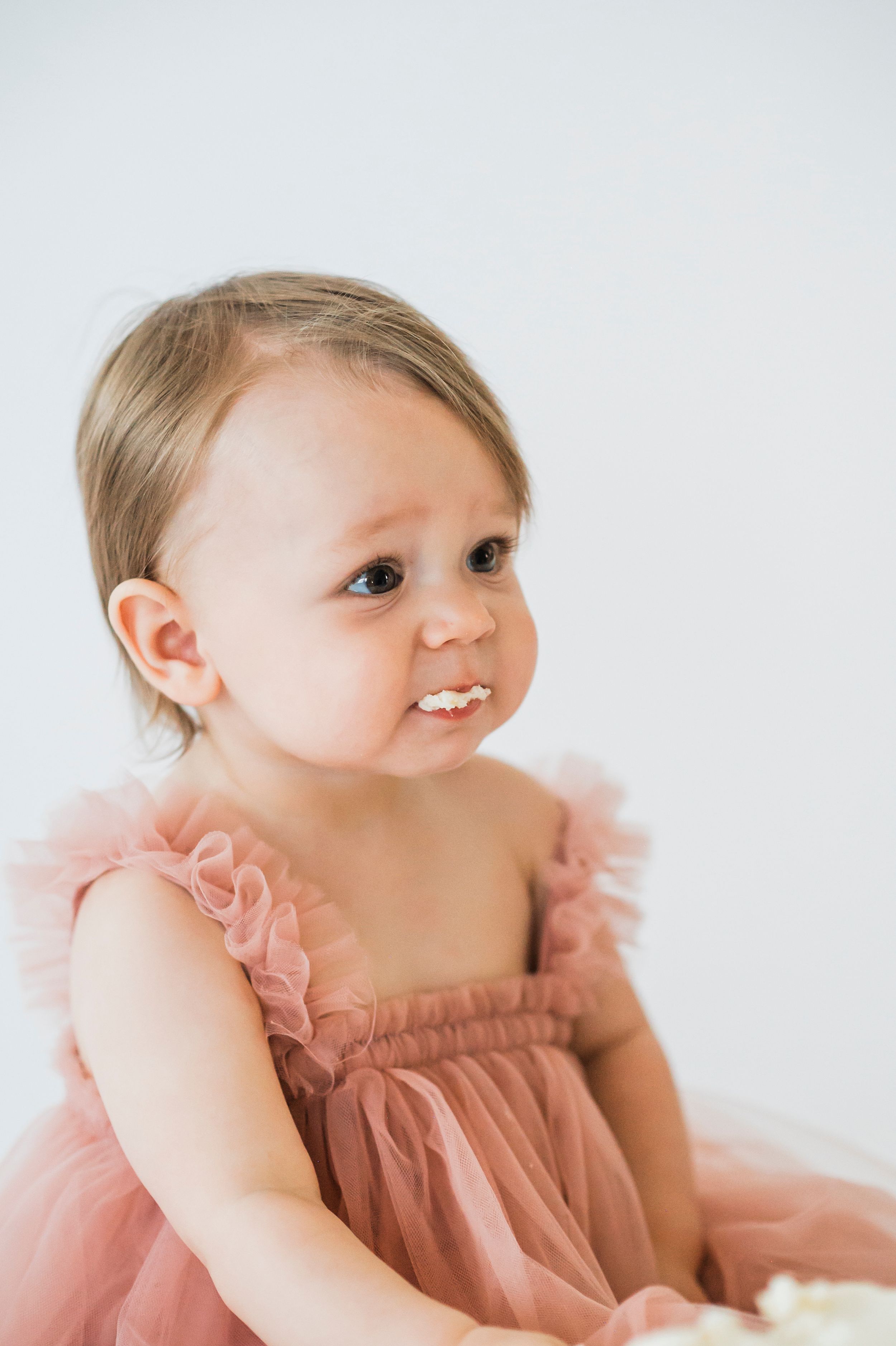 A toddler wearing a dusty pink tulle dress smiles while seated against a white background.