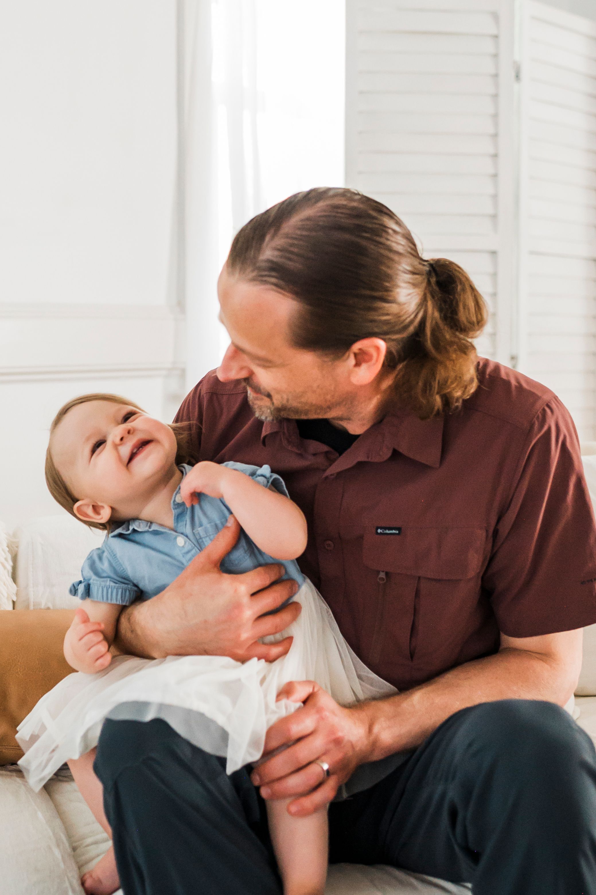 A parent in a brown shirt affectionately plays with a laughing baby wearing blue clothes while sitting on a couch.