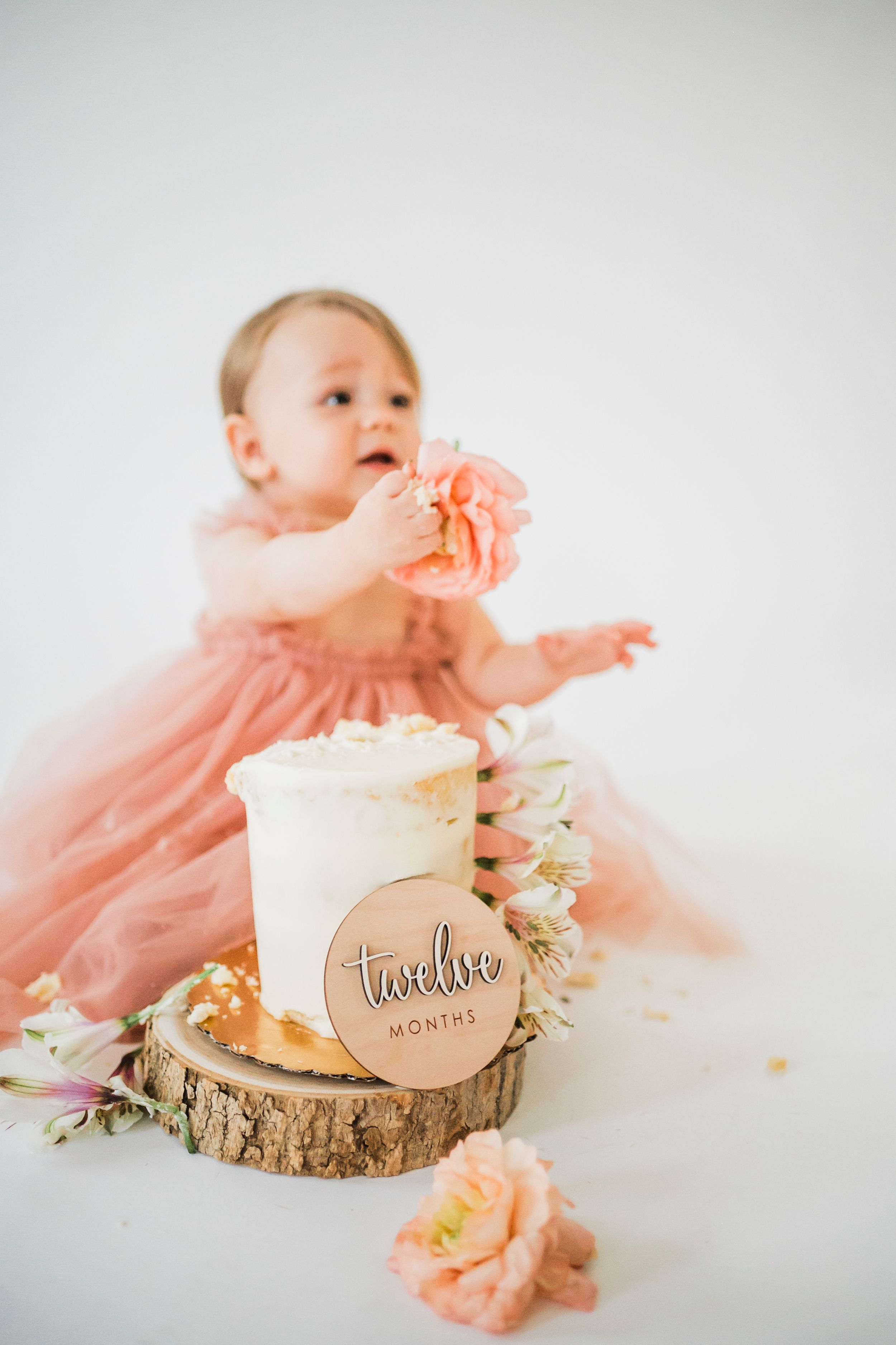 Baby in pink dress reaching for decorated cake with peach flowers on rustic wood slice.