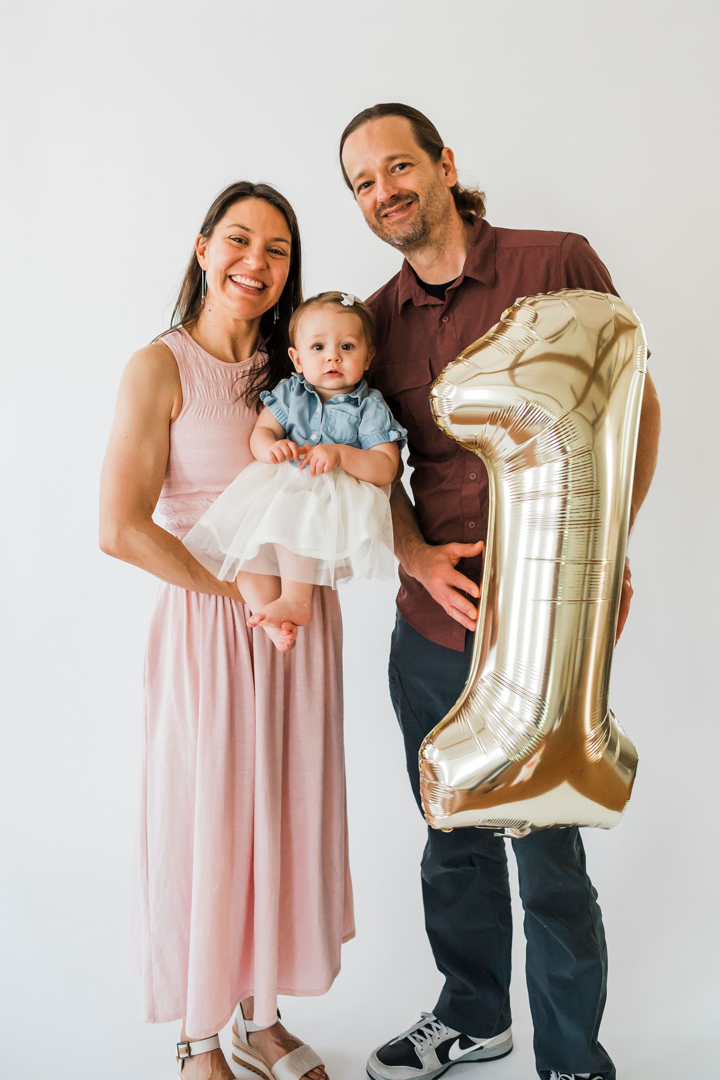 Family celebrates baby's first birthday with gold number one balloon in minimalist white studio setting.