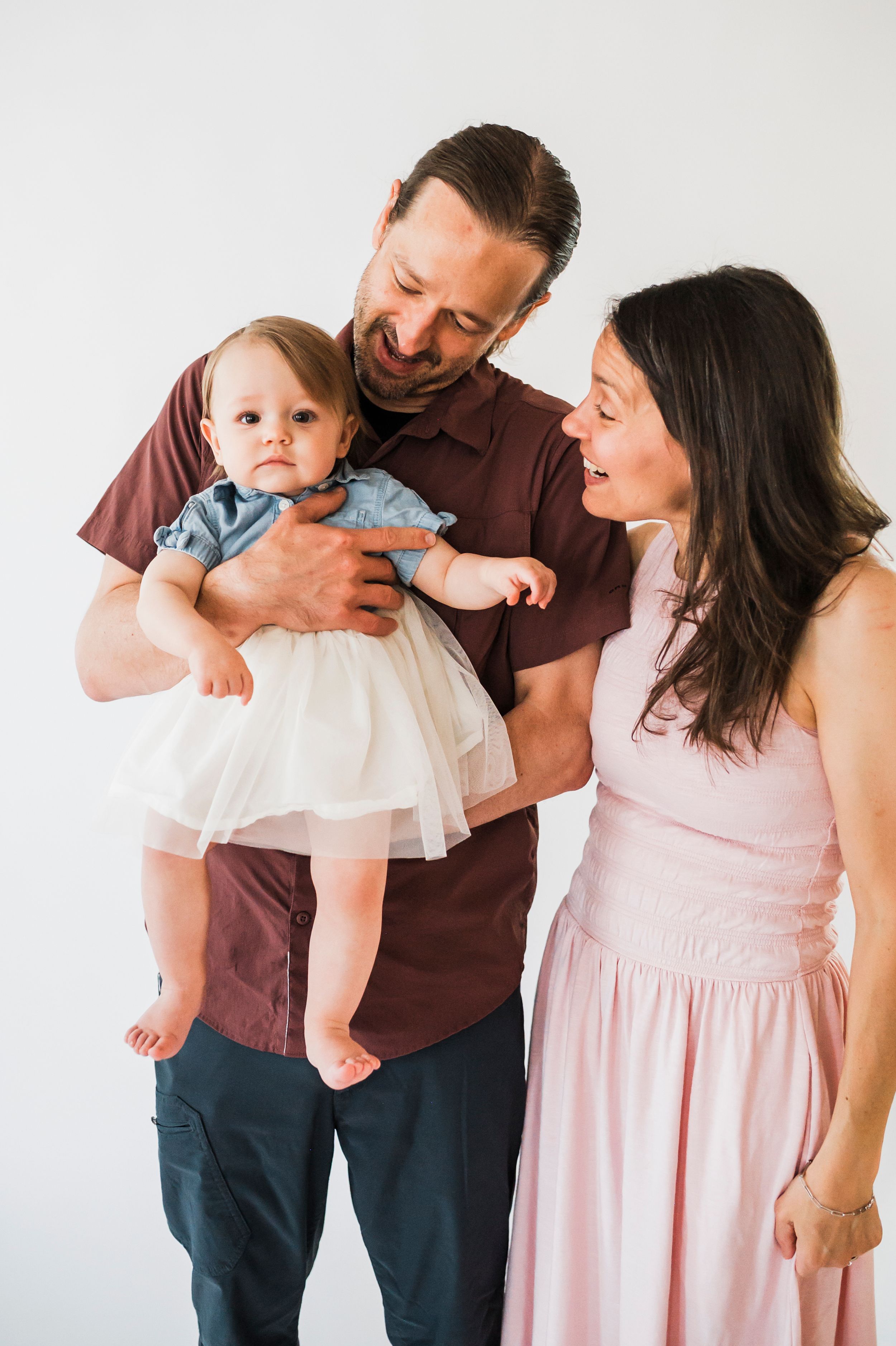 Family portrait against white backdrop with a baby in a tulle dress while parents look lovingly at their child.