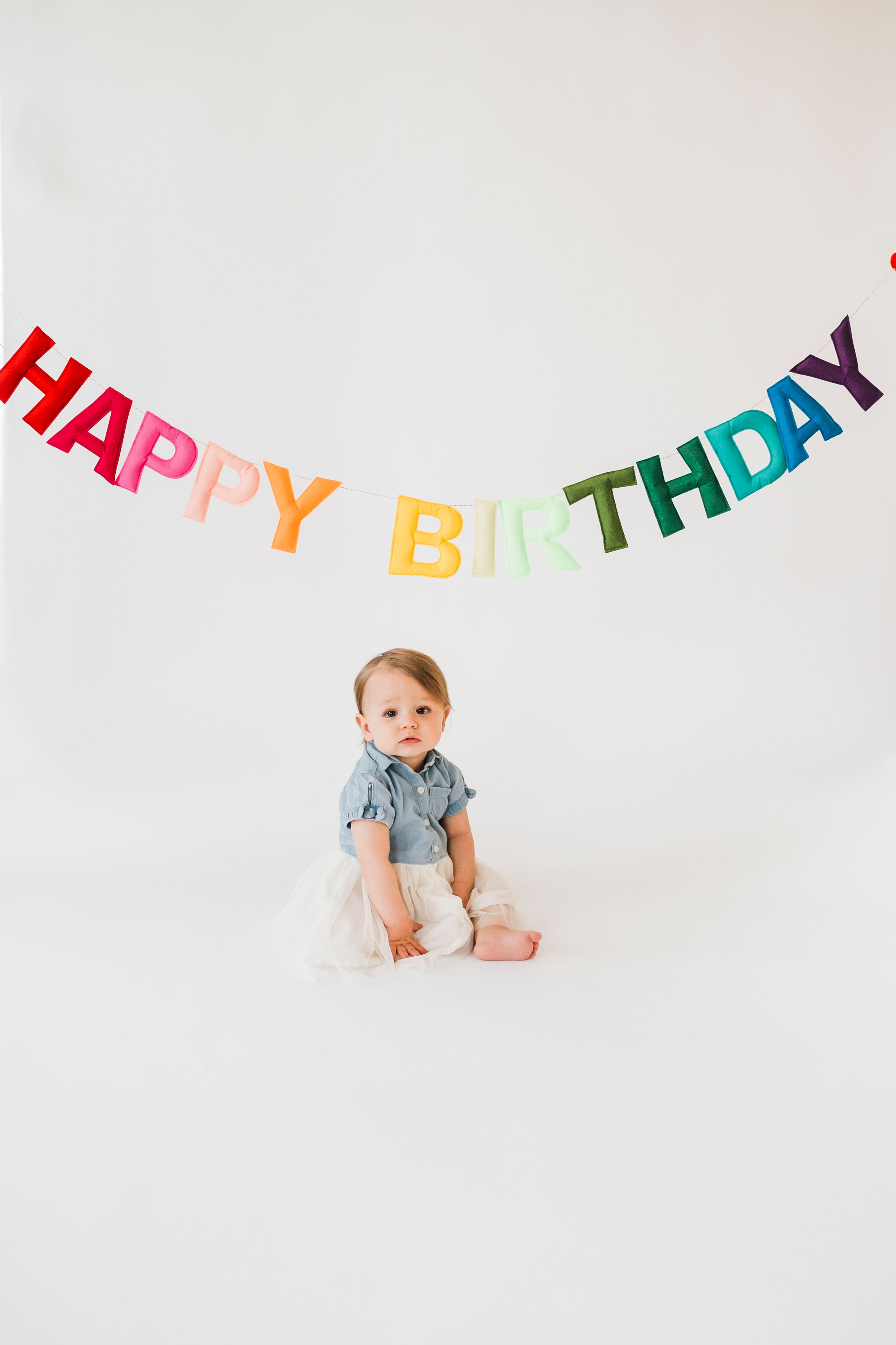 Small child sits beneath colorful rainbow Happy Birthday banner on white backdrop during photo session.