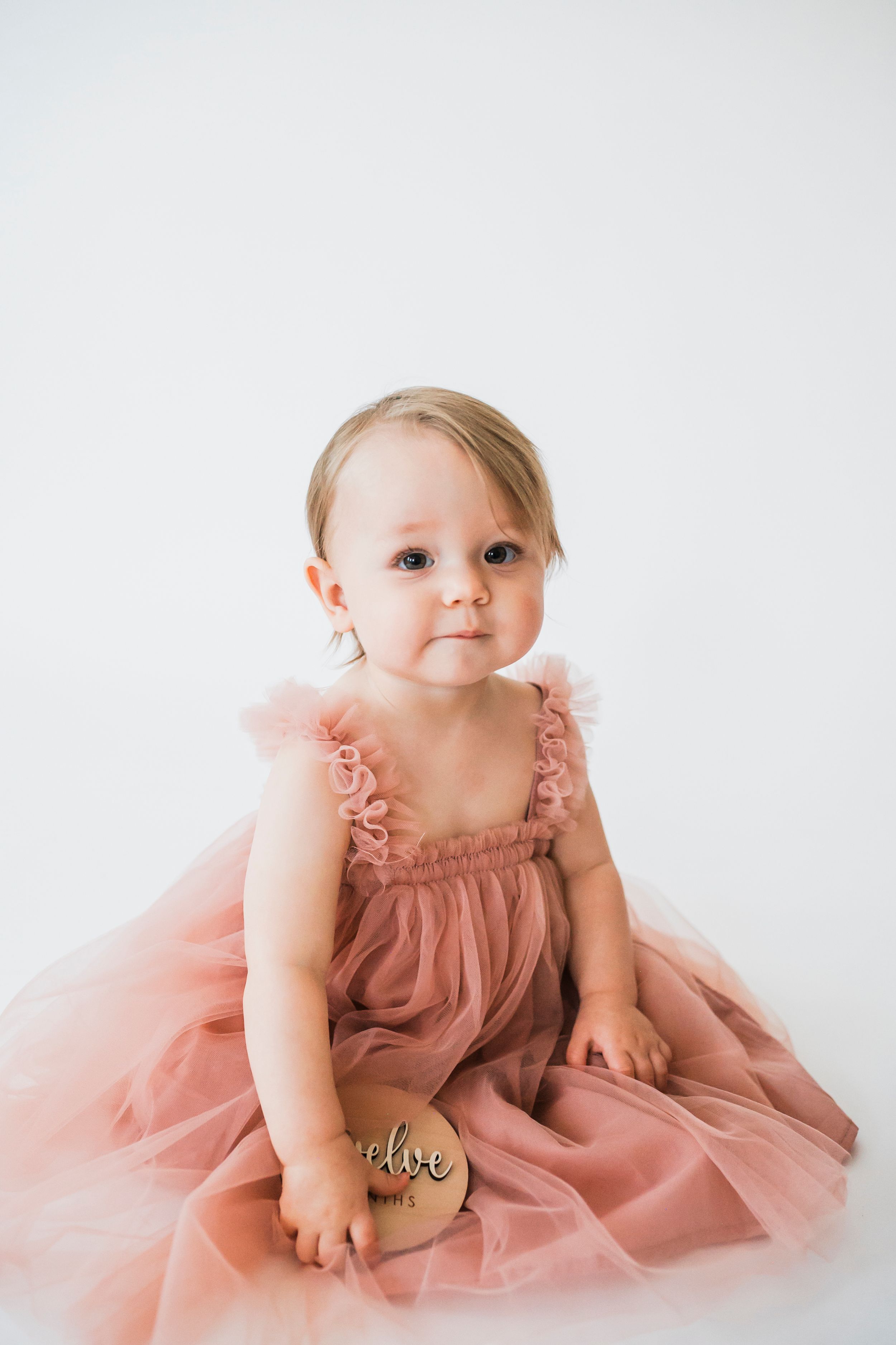 A baby wearing a pink tulle dress sits on a white background while holding a wooden block.