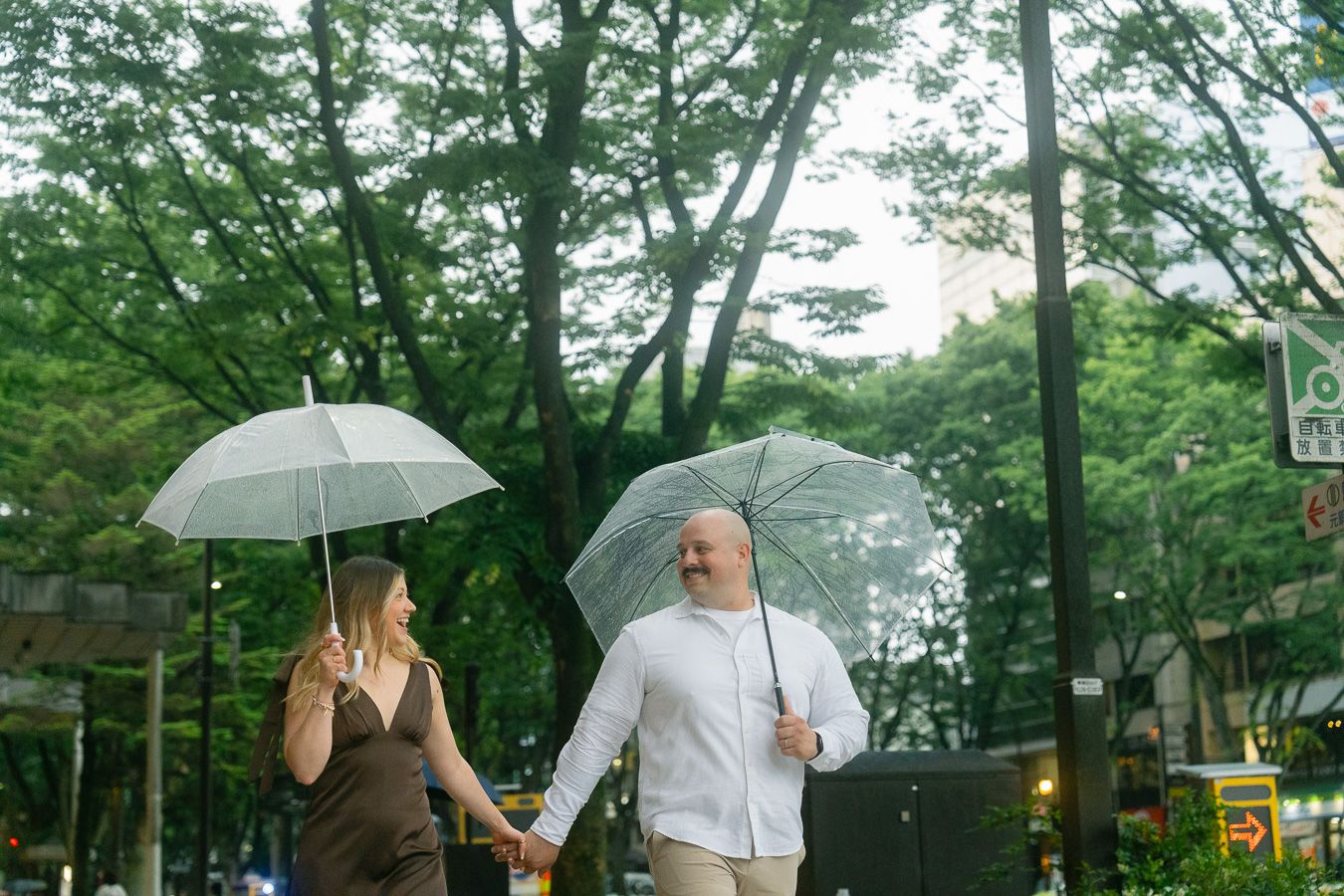 Couple holding hands and walking with clear umbrellas in the rain near lush green trees on a city street.