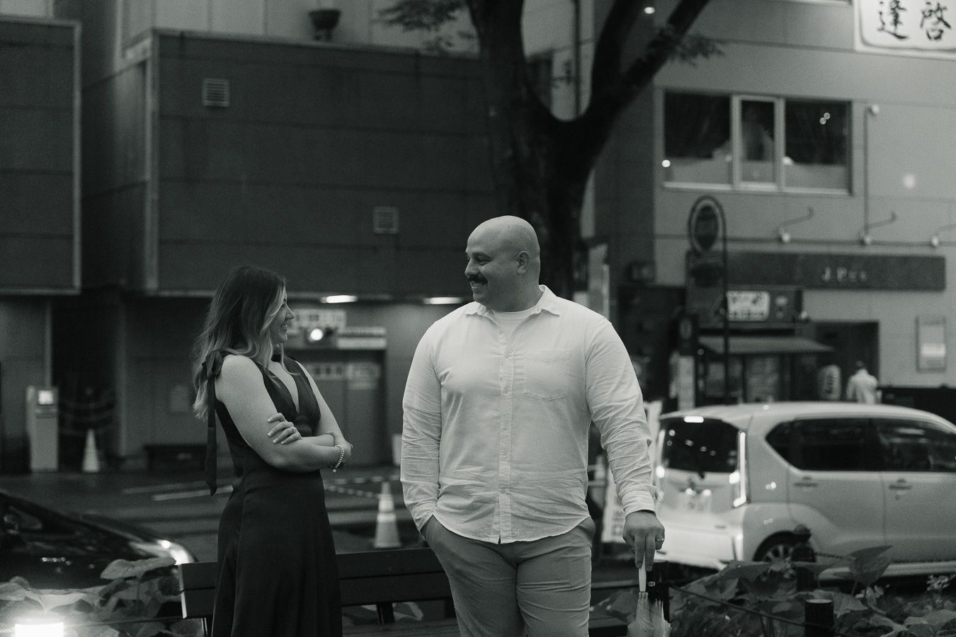 Black and white photo of a bald man and woman conversing on a city street at night.