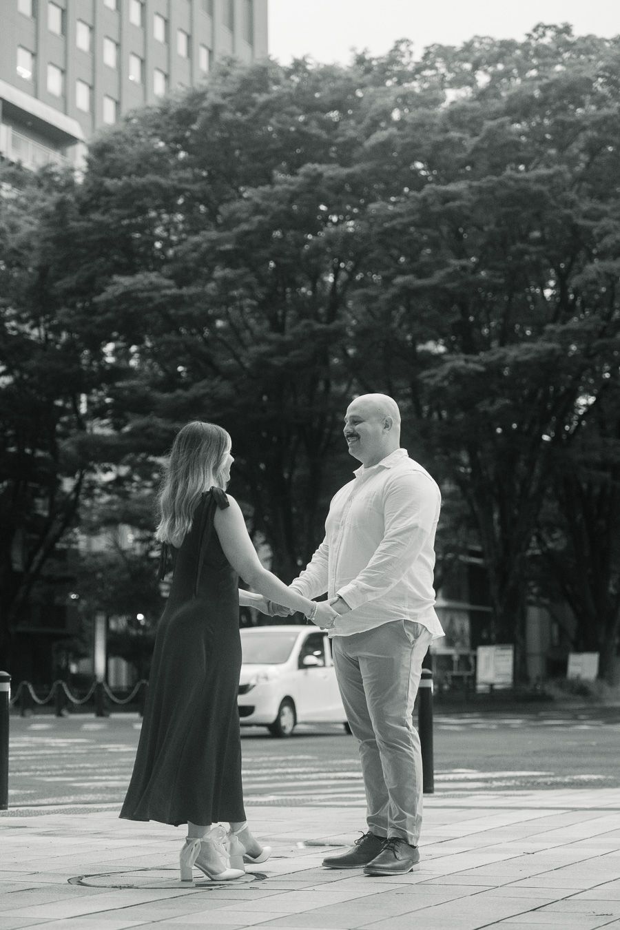Black and white photo of a couple holding hands and smiling on an urban plaza with trees and buildings behind them.