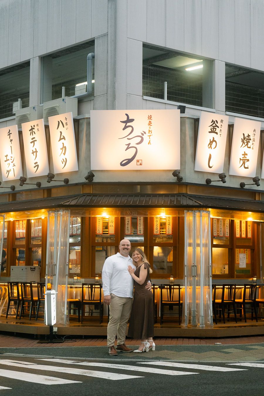 Couple posing together in front of a warmly lit traditional Japanese restaurant with lantern signs at dusk.