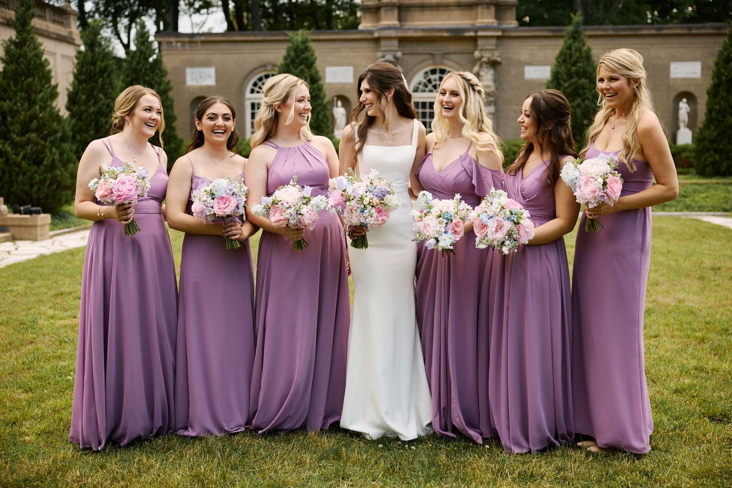 A bridal party in purple bridesmaid dresses poses together outdoors on a grassy lawn.
