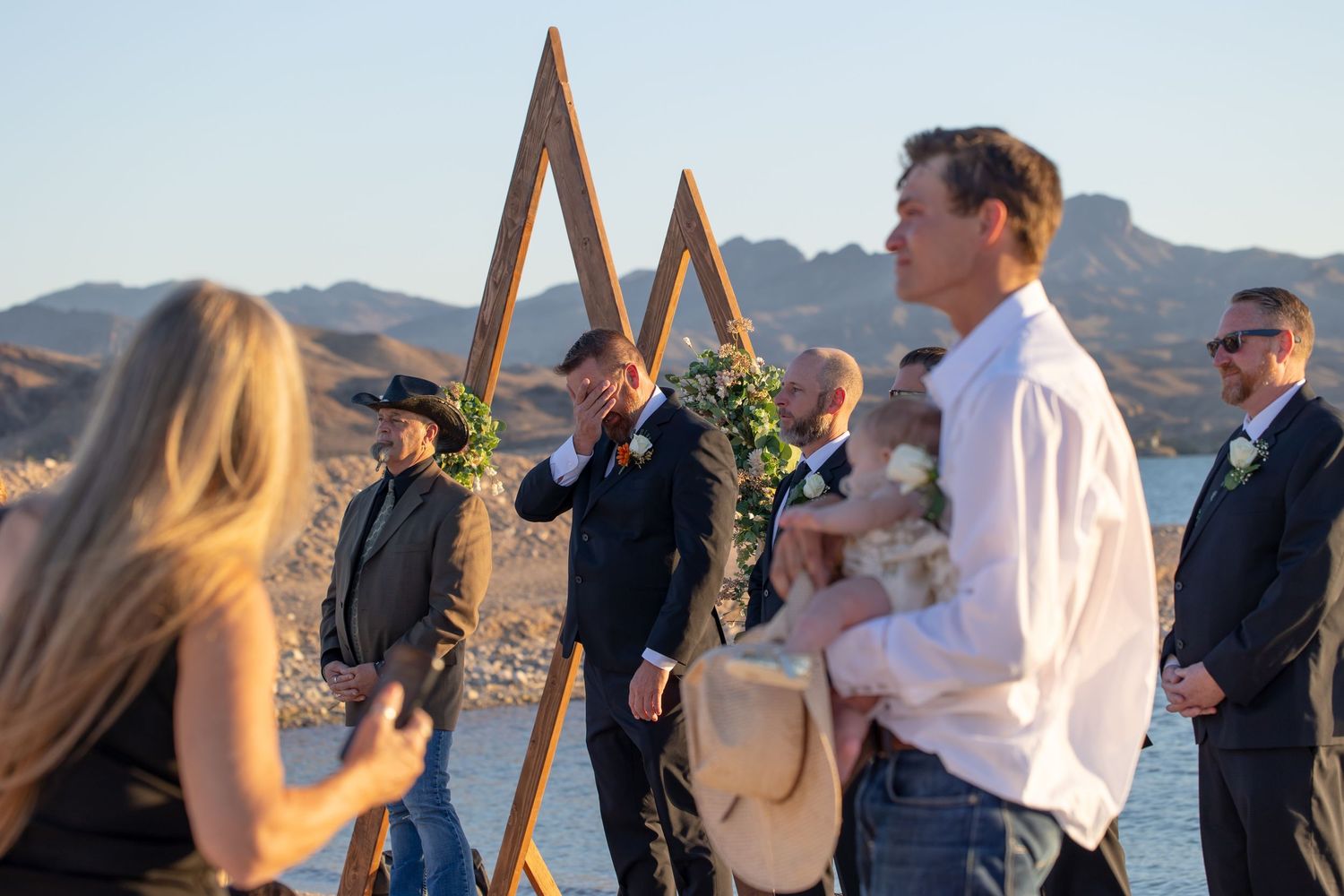 People gather at an outdoor desert wedding ceremony setup with wooden triangle arches in the mountains at sunset.