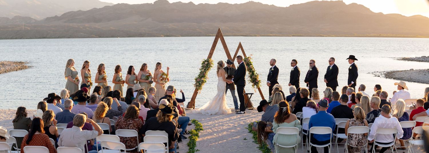 Panoramic view of a beach wedding ceremony setup and guests gathering along a tropical shoreline during sunset.