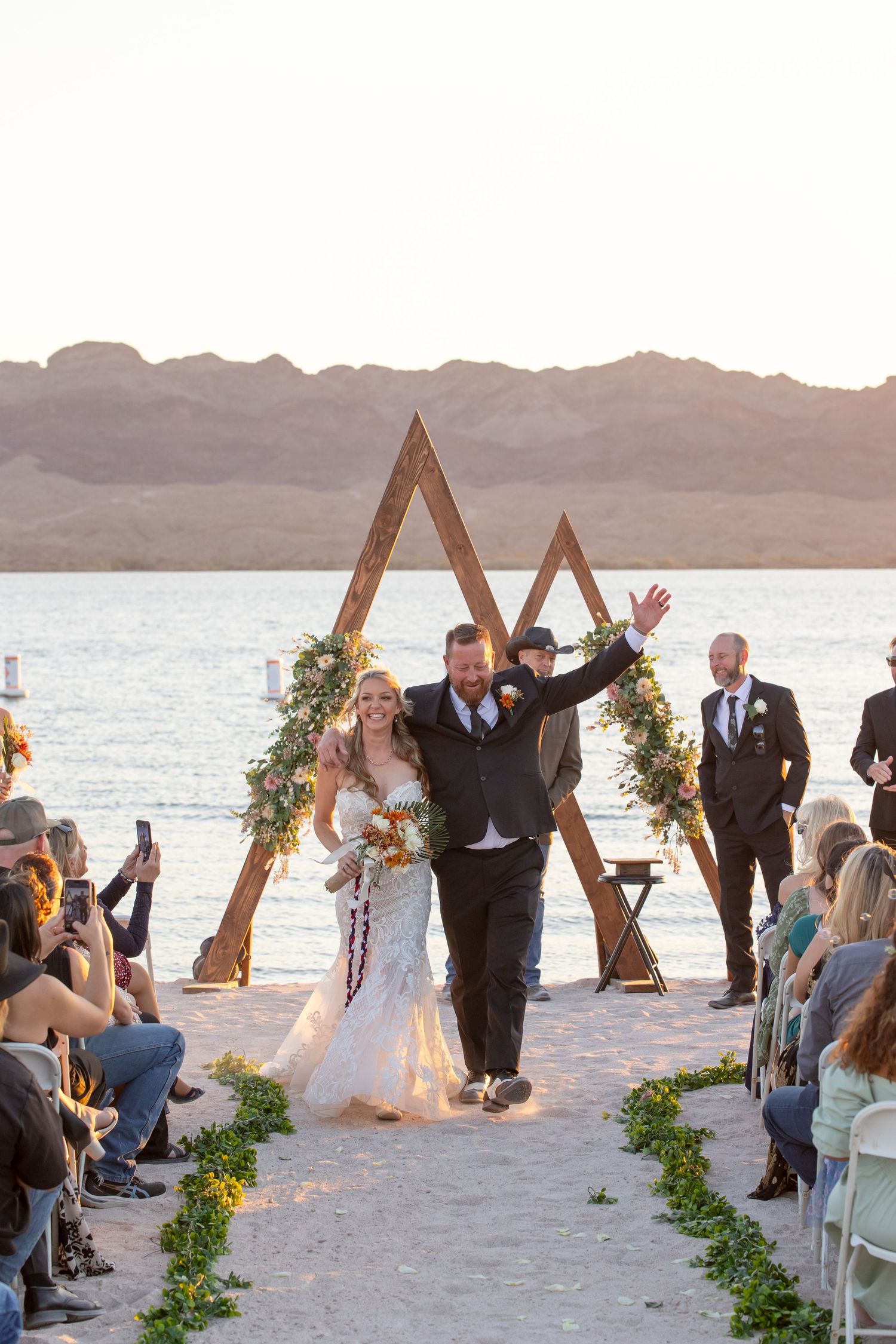Beachfront wedding ceremony with wooden triangle arch and greenery-lined aisle at sunset with lake and mountain views.