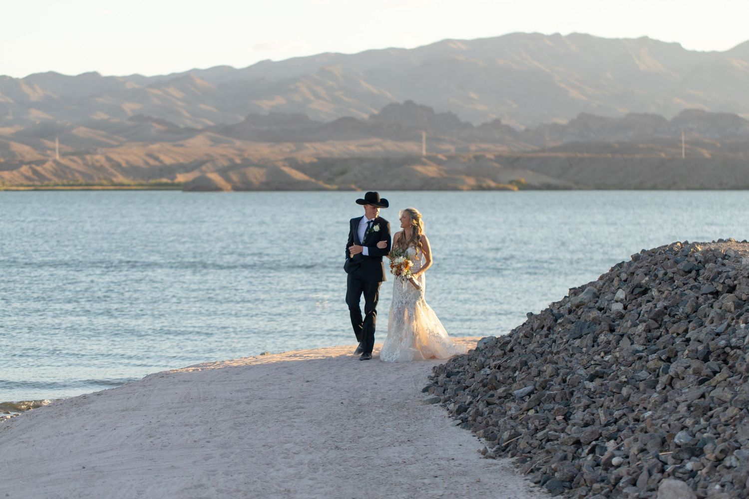 A couple walks along a rocky path beside a scenic lake with mountains in the background at sunset.