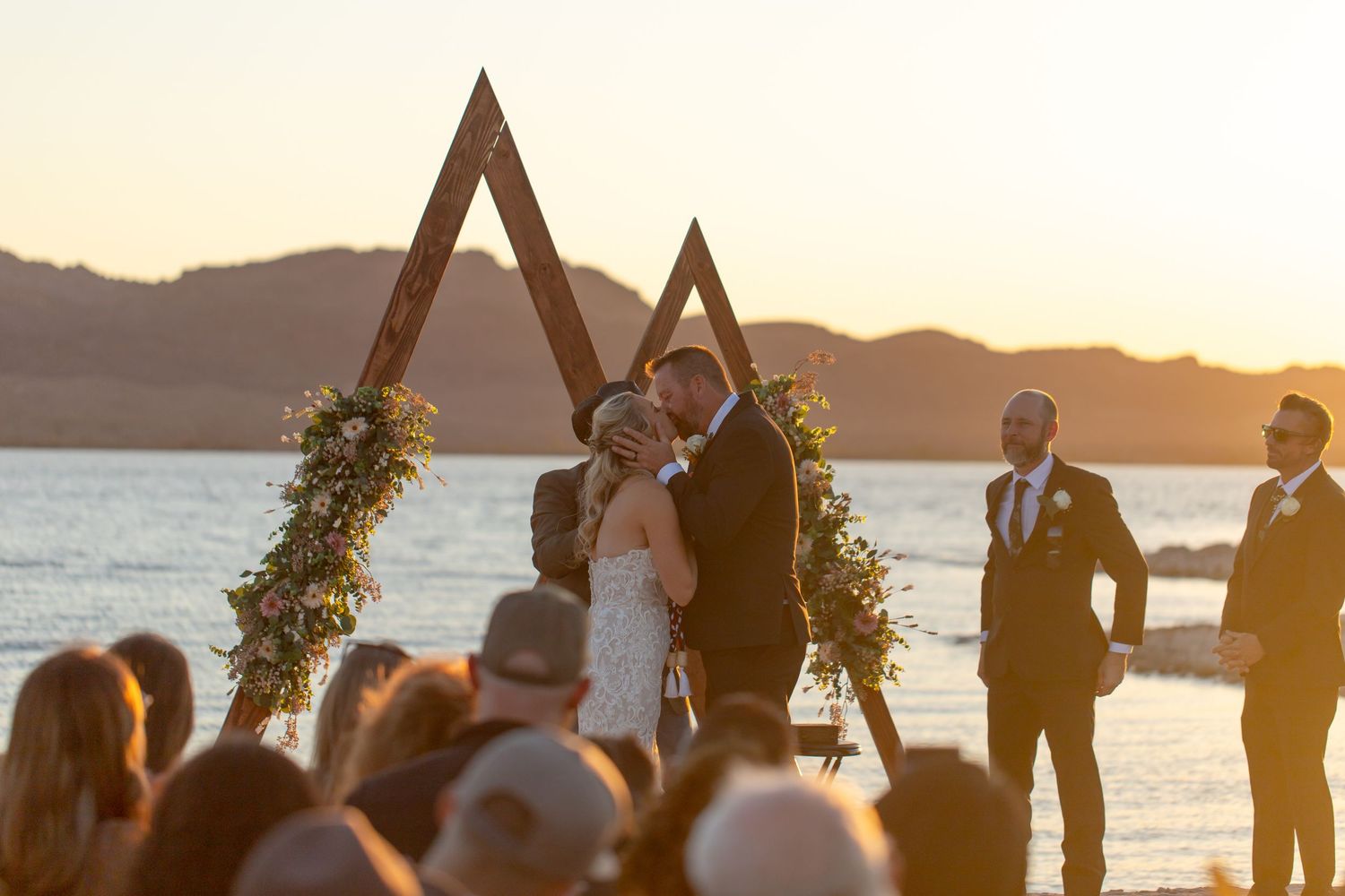 Outdoor waterfront wedding ceremony at sunset with a triangular wooden arch decorated with greenery and flowers.
