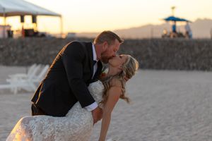 A dipping kiss shared between a couple in formal attire on a scenic beach at sunset.