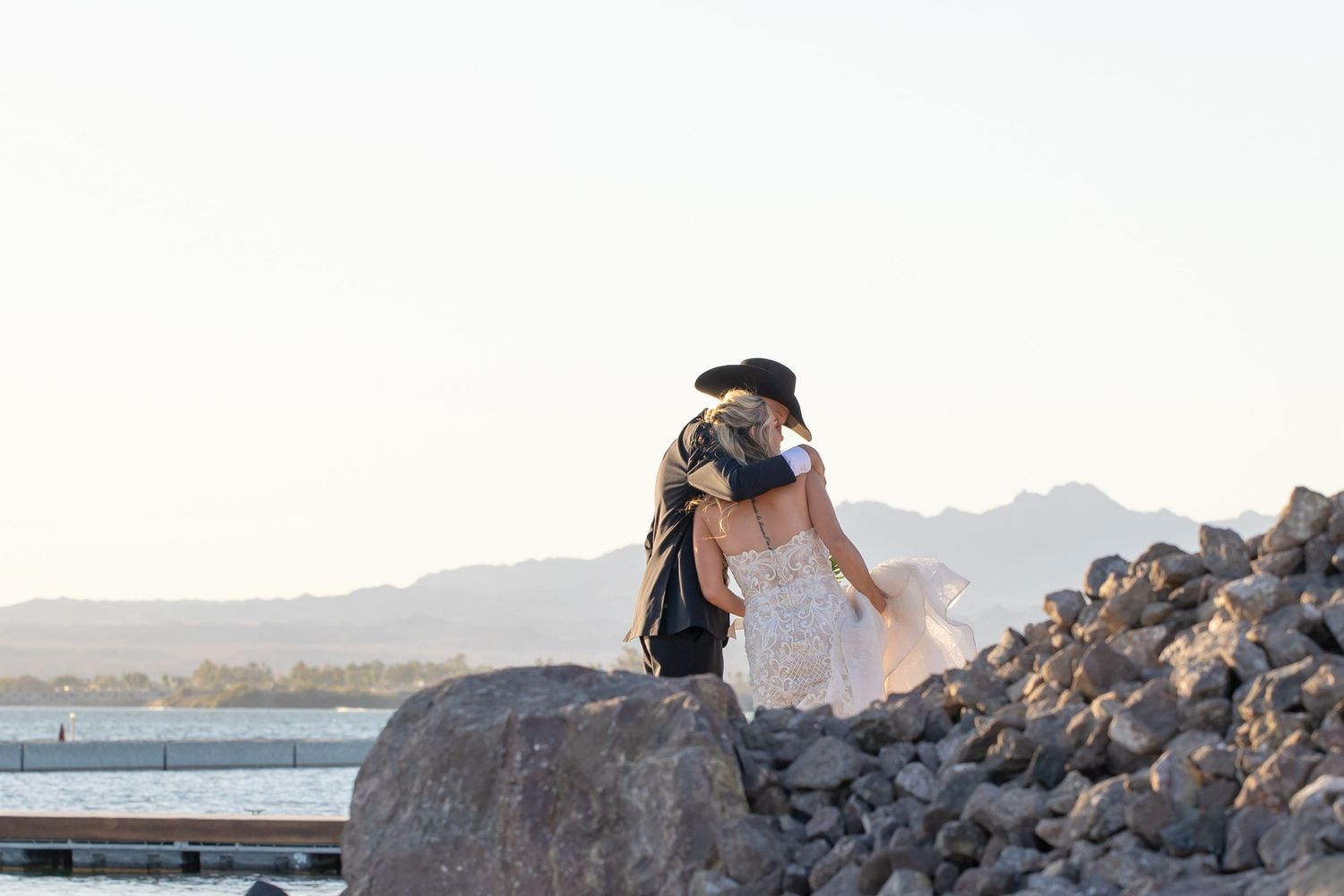 Two people embrace while sitting on coastal rocks near the ocean at sunset.