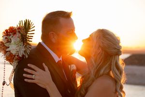 Romantic golden hour wedding portrait with silhouetted couple embracing against sunset sky.