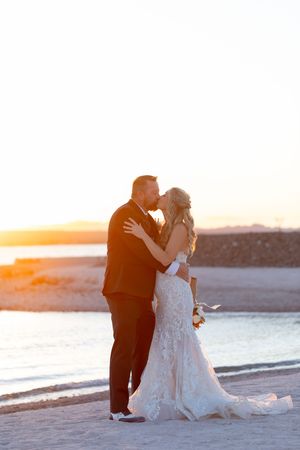 A romantic couple embraces on a beach at sunset, with the bride wearing a lace wedding gown and groom in a black suit.