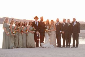 Wedding party members in light colored suits and dresses pose together at sunset on the beach for formal photos.