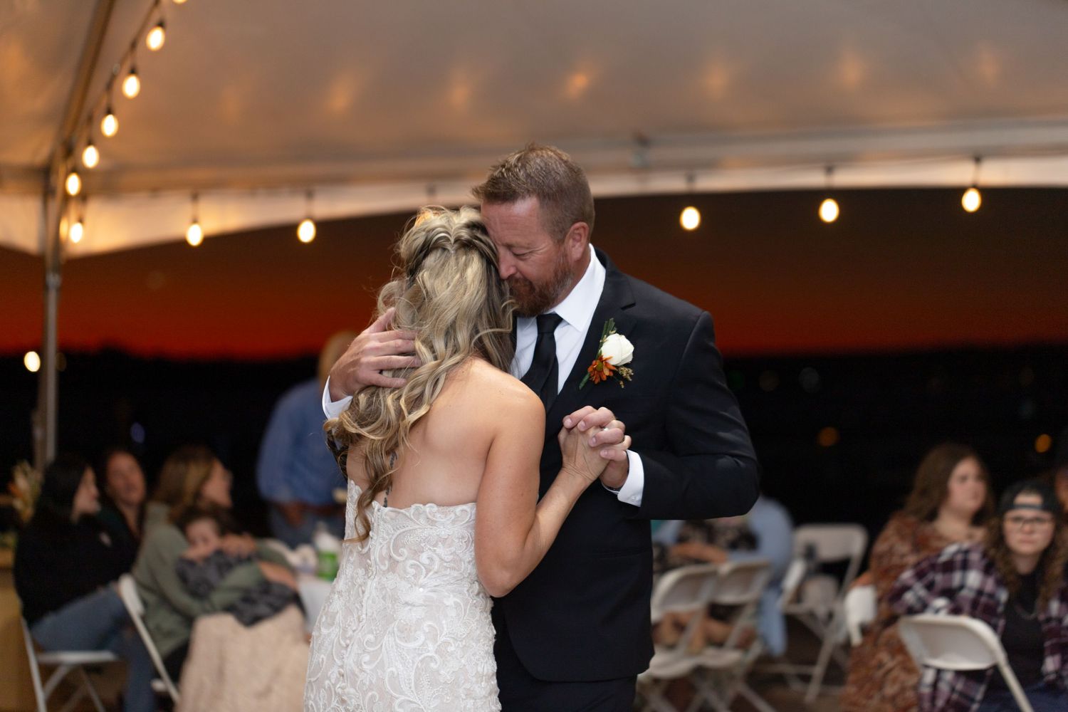 A couple dances together beneath string lights at their wedding reception with guests watching in the background.