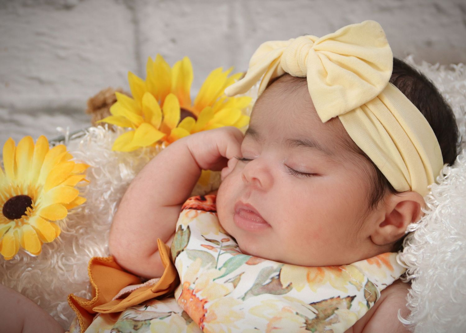 Sleeping baby with yellow bow headband surrounded by sunflowers on white textured background.