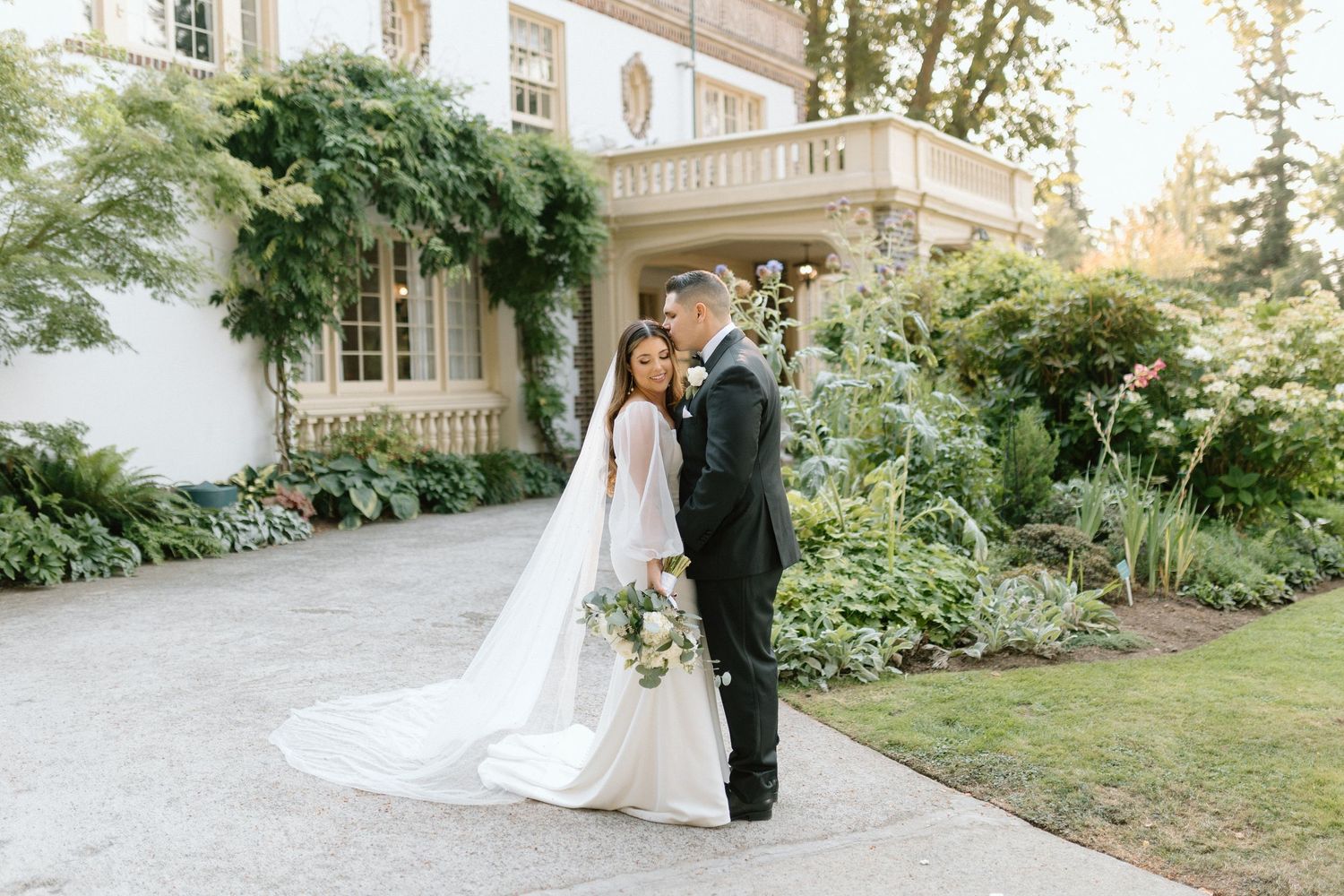 A couple in wedding attire stands together on a curved driveway outside an elegant mansion with lush gardens.