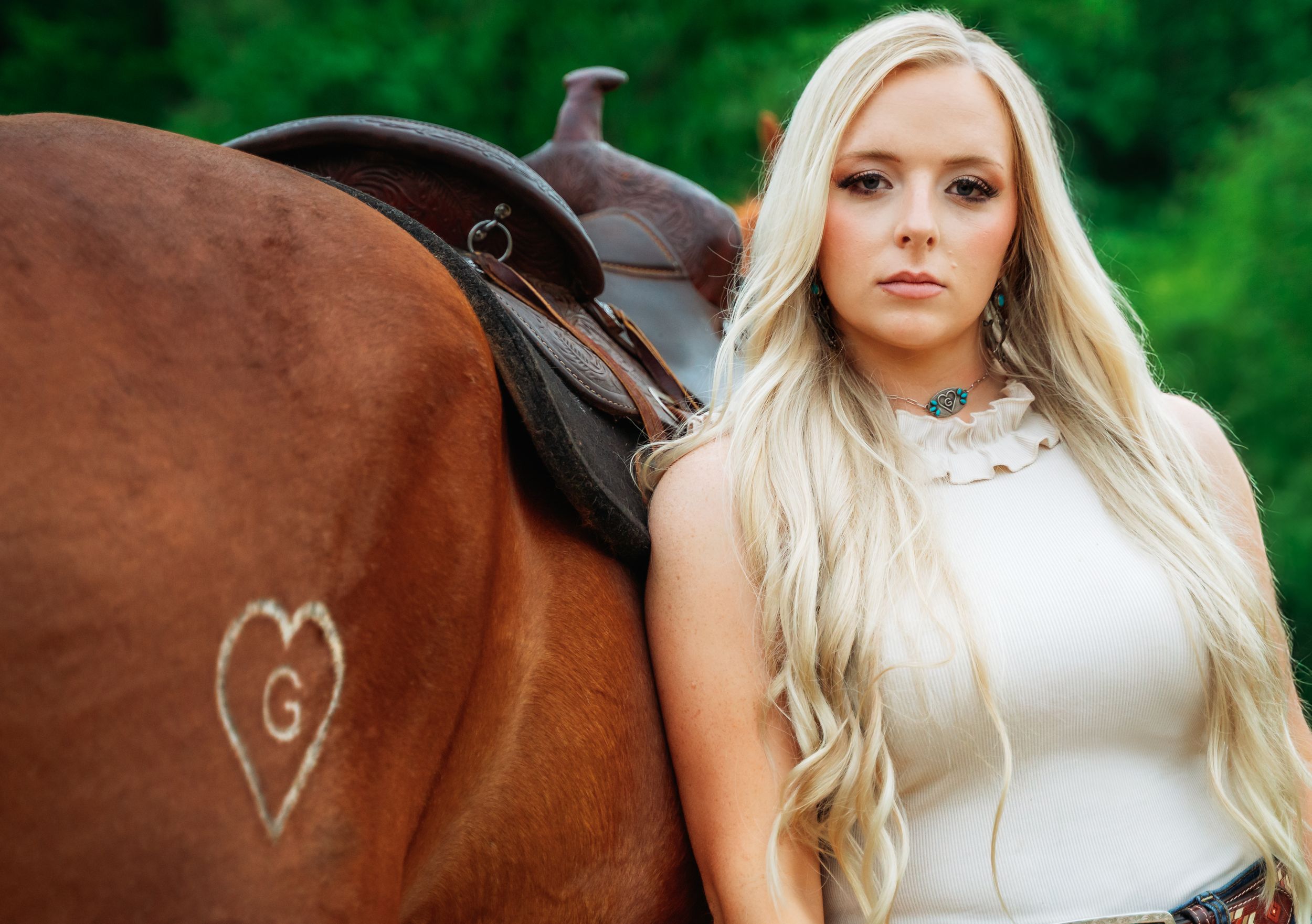 A brown horse with a heart symbol on its side stands next to a white saddle in a green outdoor setting.