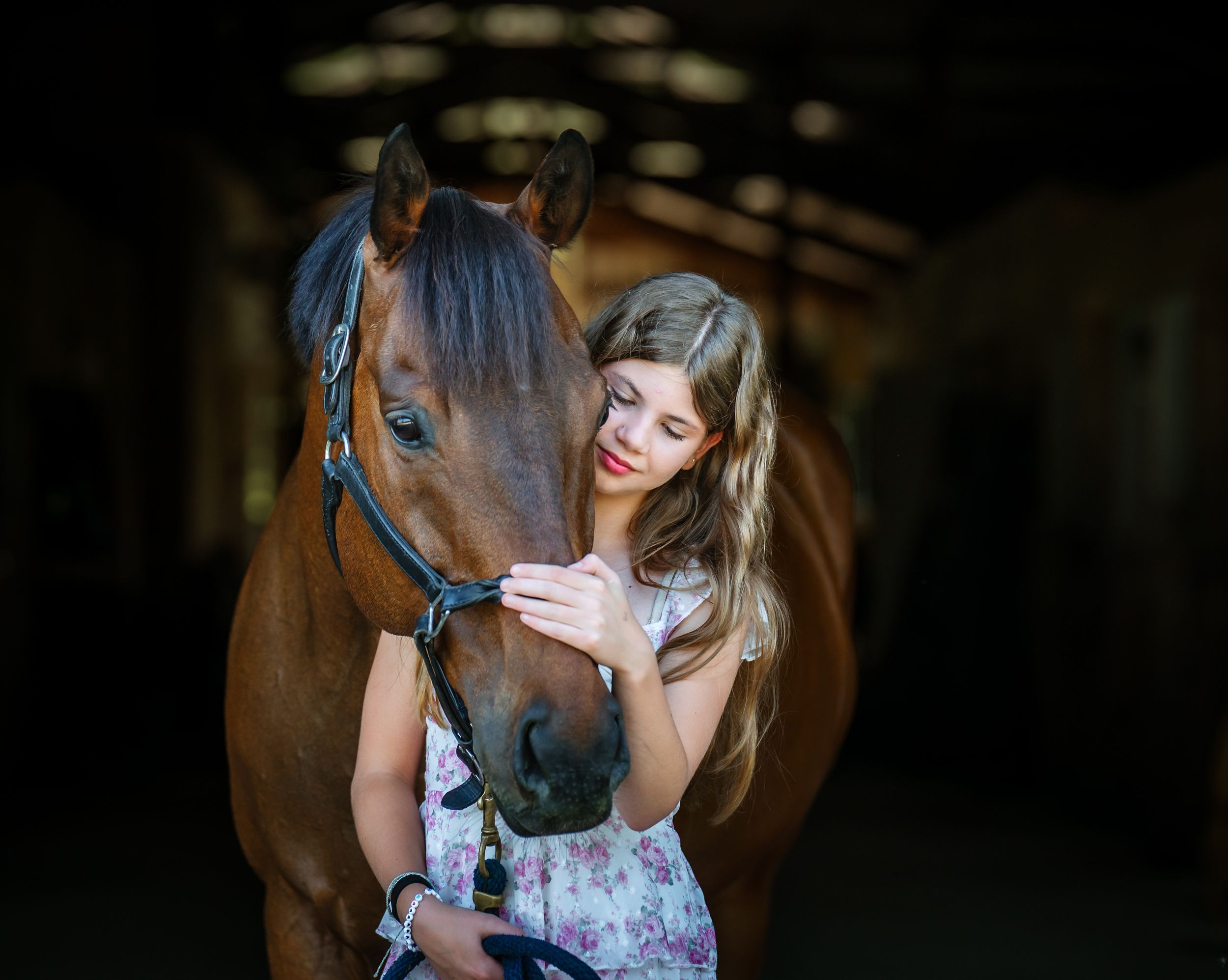 Young woman in floral dress gently pet a brown horse in a dark stable as they share a tender moment together.
