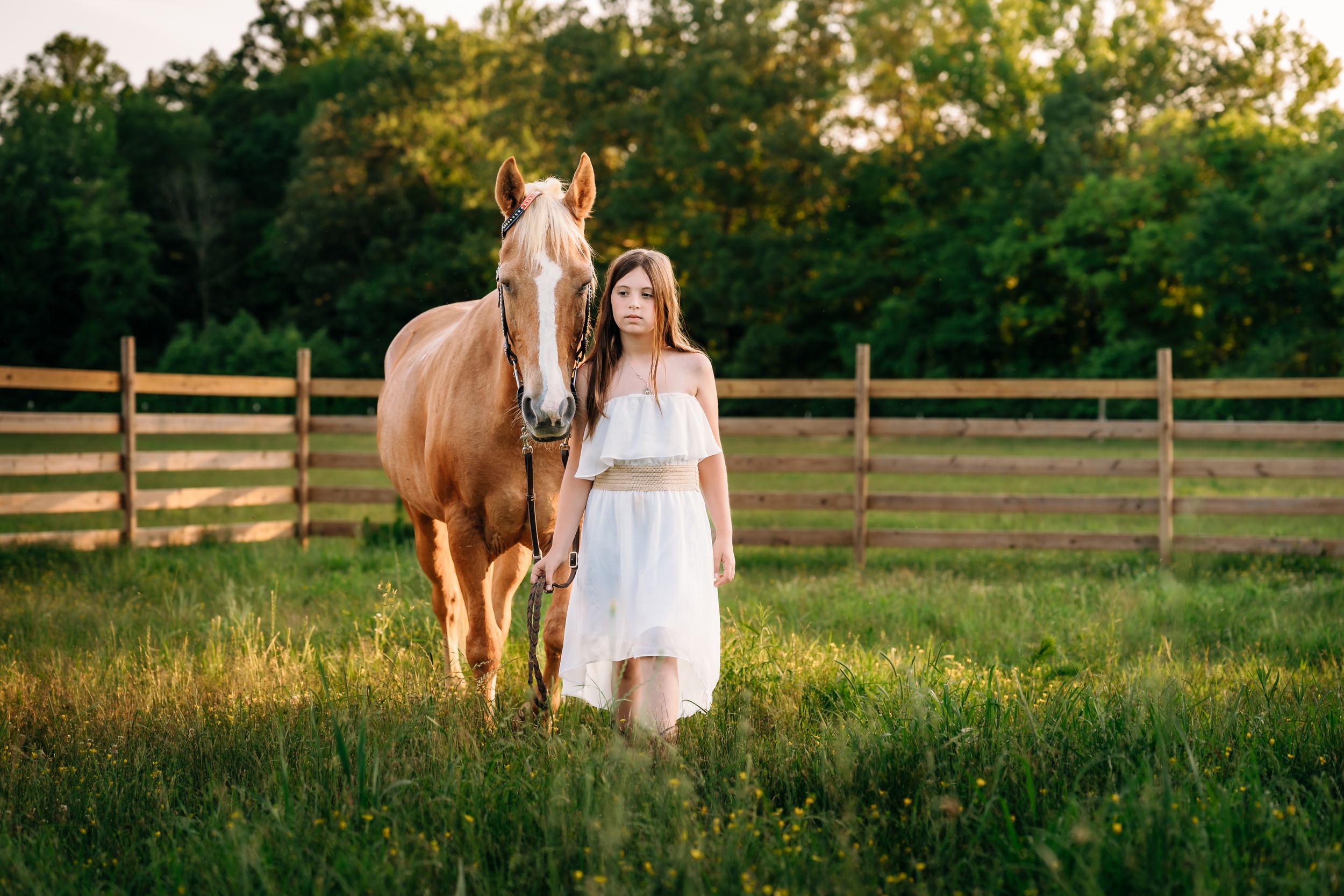 A woman in a white dress stands beside a brown horse in a fenced pasture during golden hour.