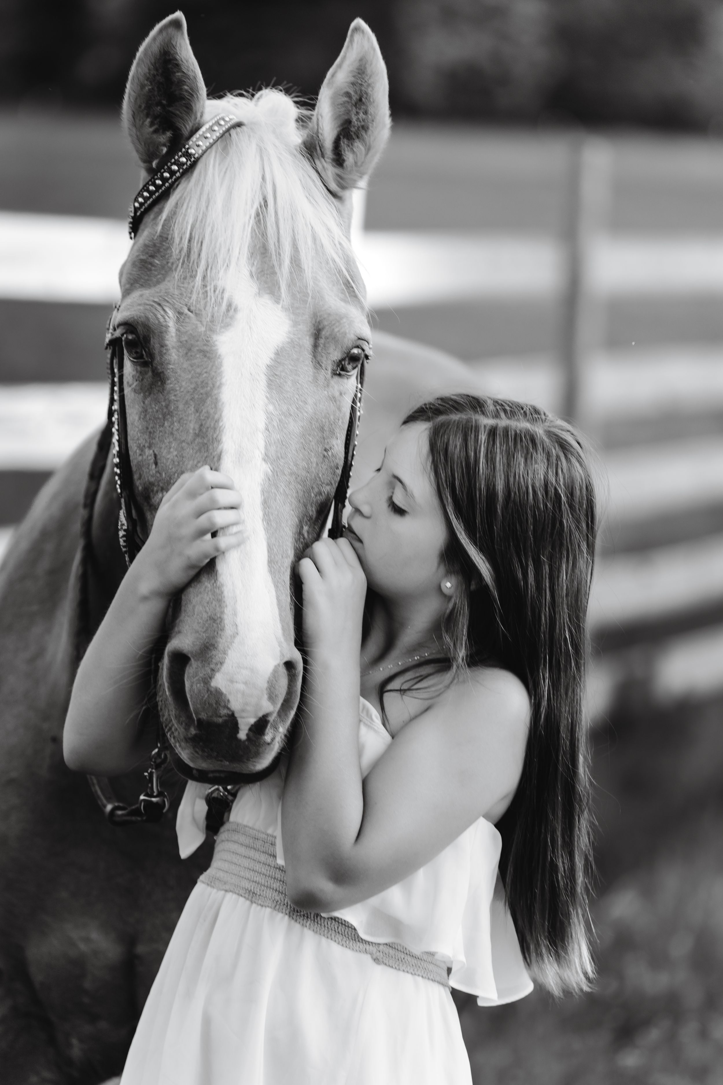 Black and white photo of a young girl in white dress kissing a light-colored horse's nose in front of a fence.