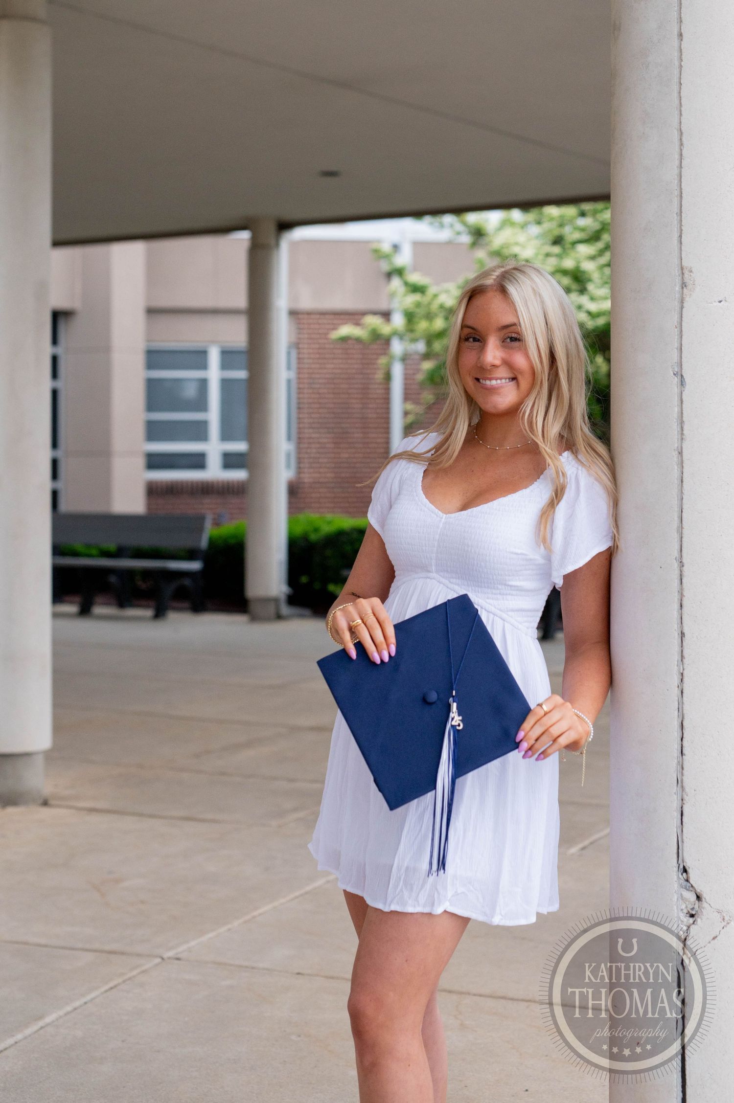 Student in white dress celebrates at graduation with blue cap and diploma in outdoor colonnade walkway.