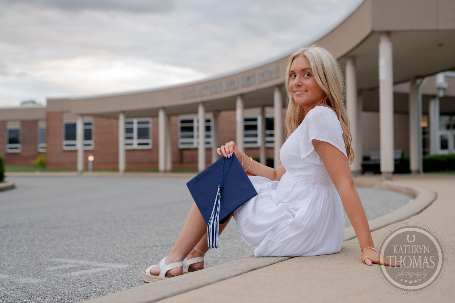 Graduating student in white dress sits with blue graduation cap outside a school building on a cloudy day.