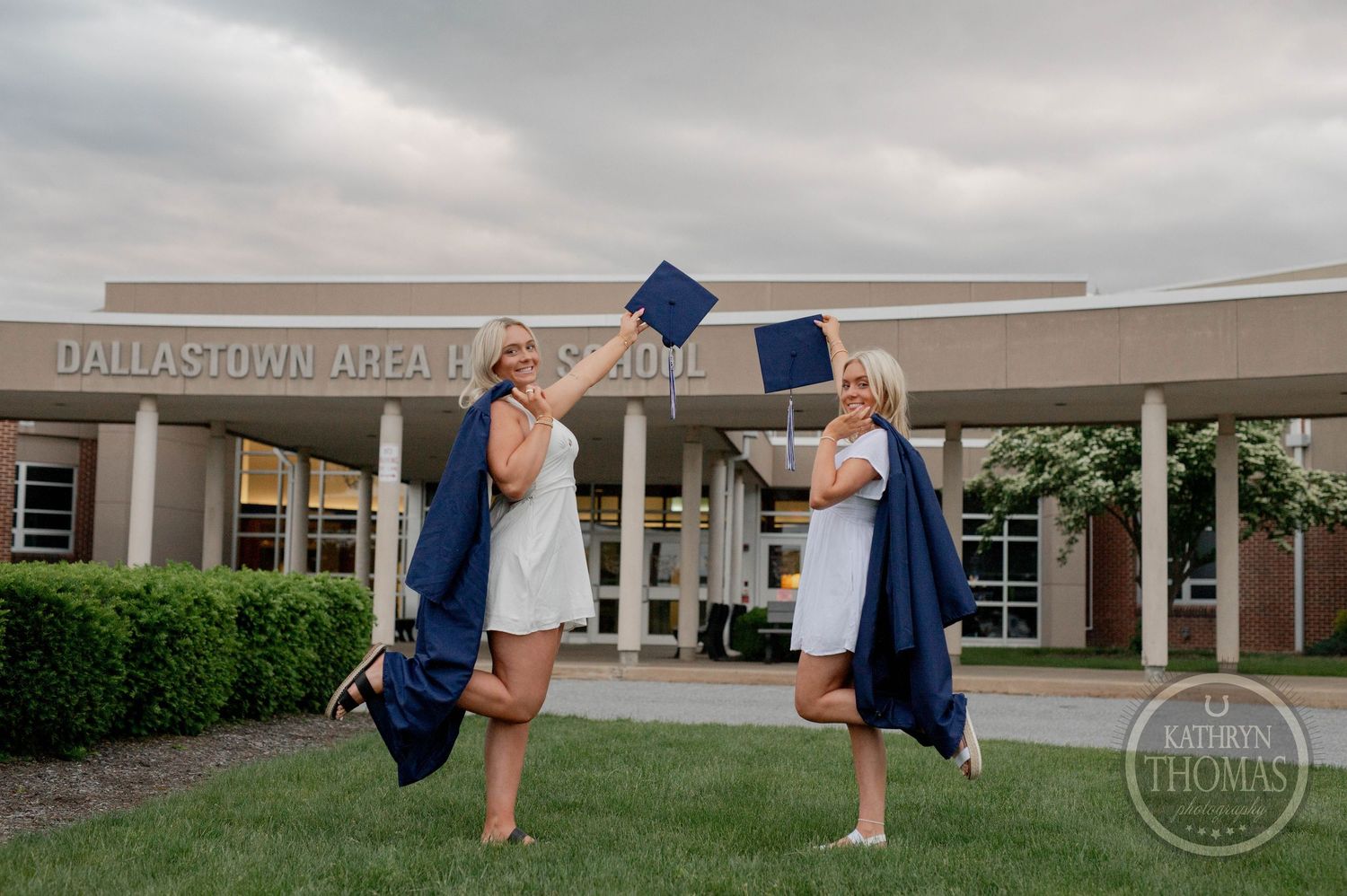 Two graduates in blue caps and gowns excitedly throw their graduation caps in front of a brick building entrance.