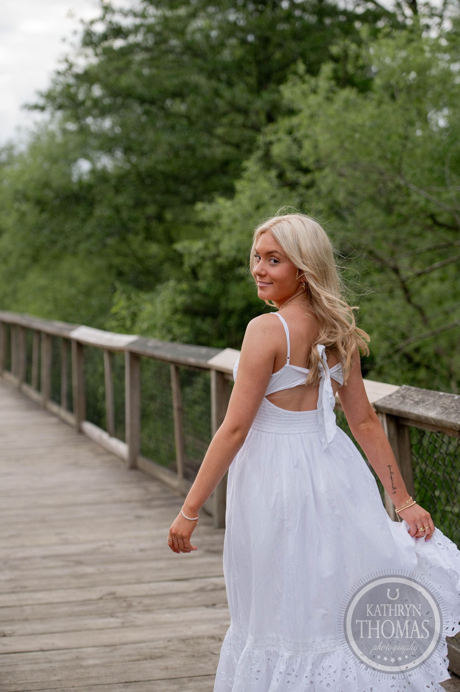Woman in flowing white dress poses on wooden boardwalk surrounded by lush green trees on summer day.