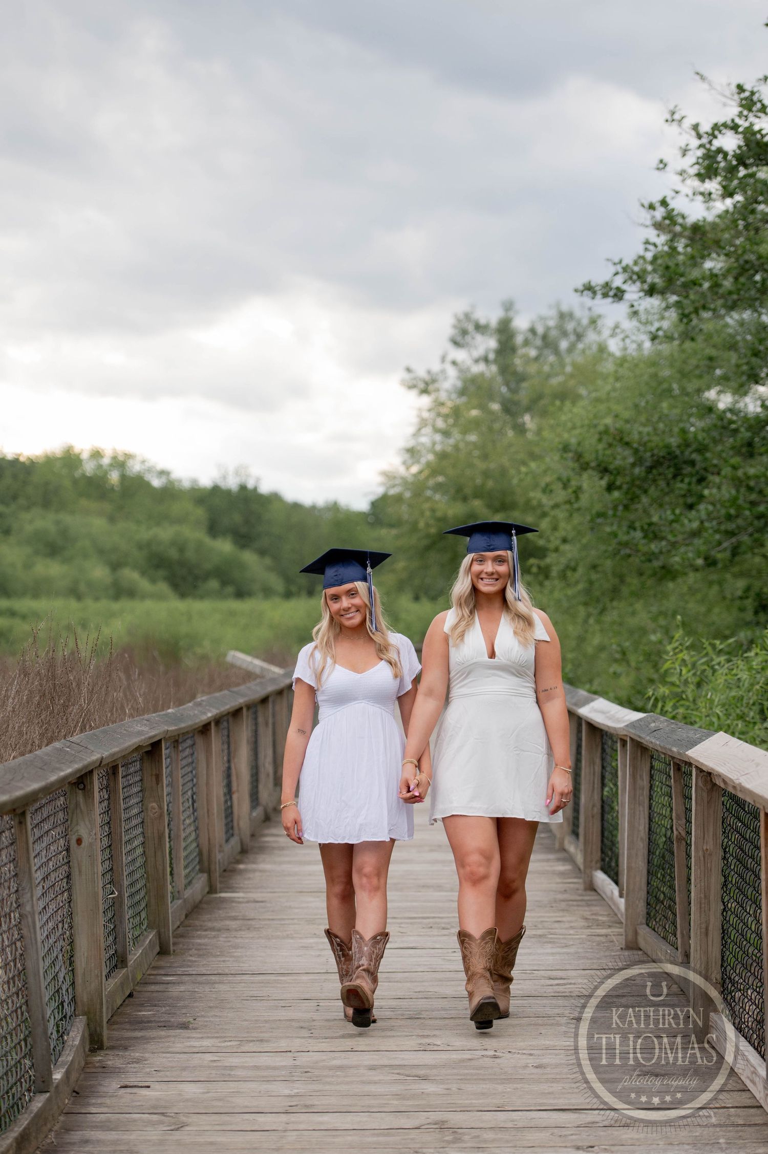 Two graduates in white dresses and cowboy boots walk hand-in-hand along a wooden boardwalk path surrounded by greenery.